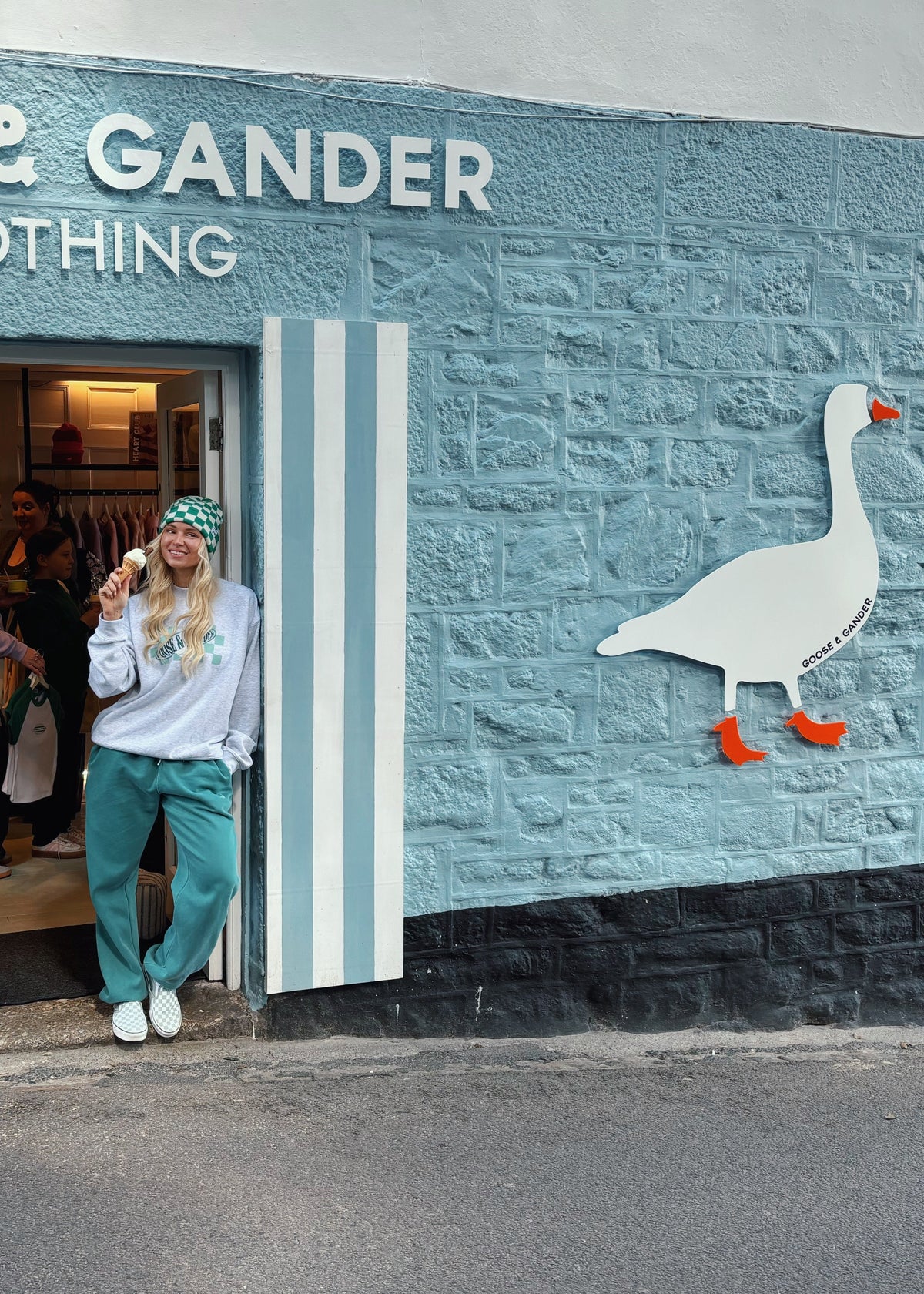 A person stands in the doorway of Goose & Gander Clothing, wearing a white sweatshirt and teal pants. The storefront features a textured light blue brick wall with white lettering, striped shutters, a large goose mural, and a giant goose sculpture on the sidewalk.